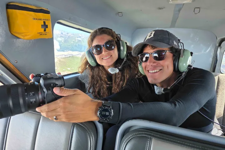 Two people in a plane wearing headsets, one holding a camera towards a window.