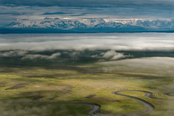 Aerial view of a winding river and grassy plains under clouds with snowy mountains in the distance.