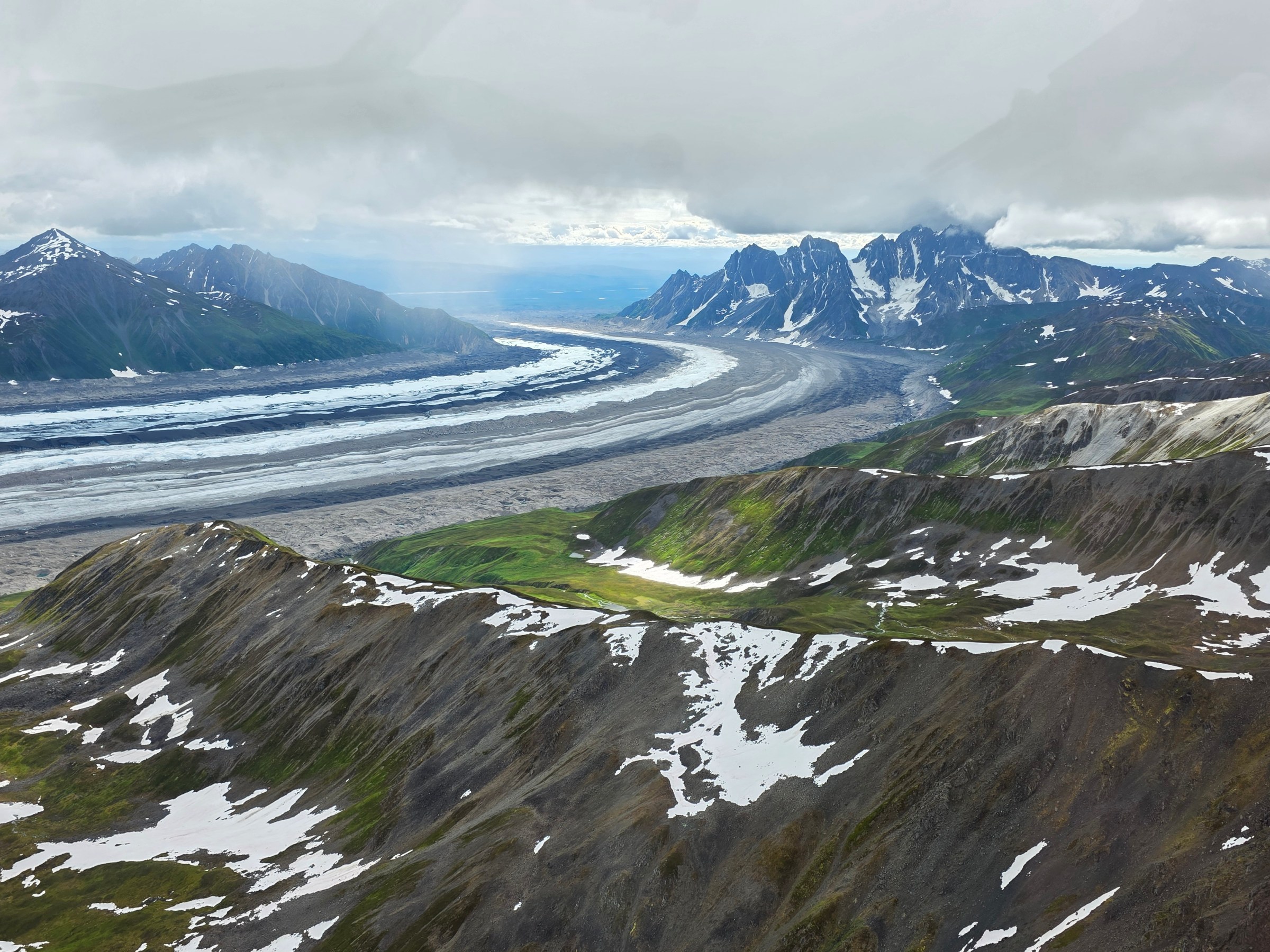 Aerial view of a mountainous landscape with snow patches and a winding glacier under cloudy skies.