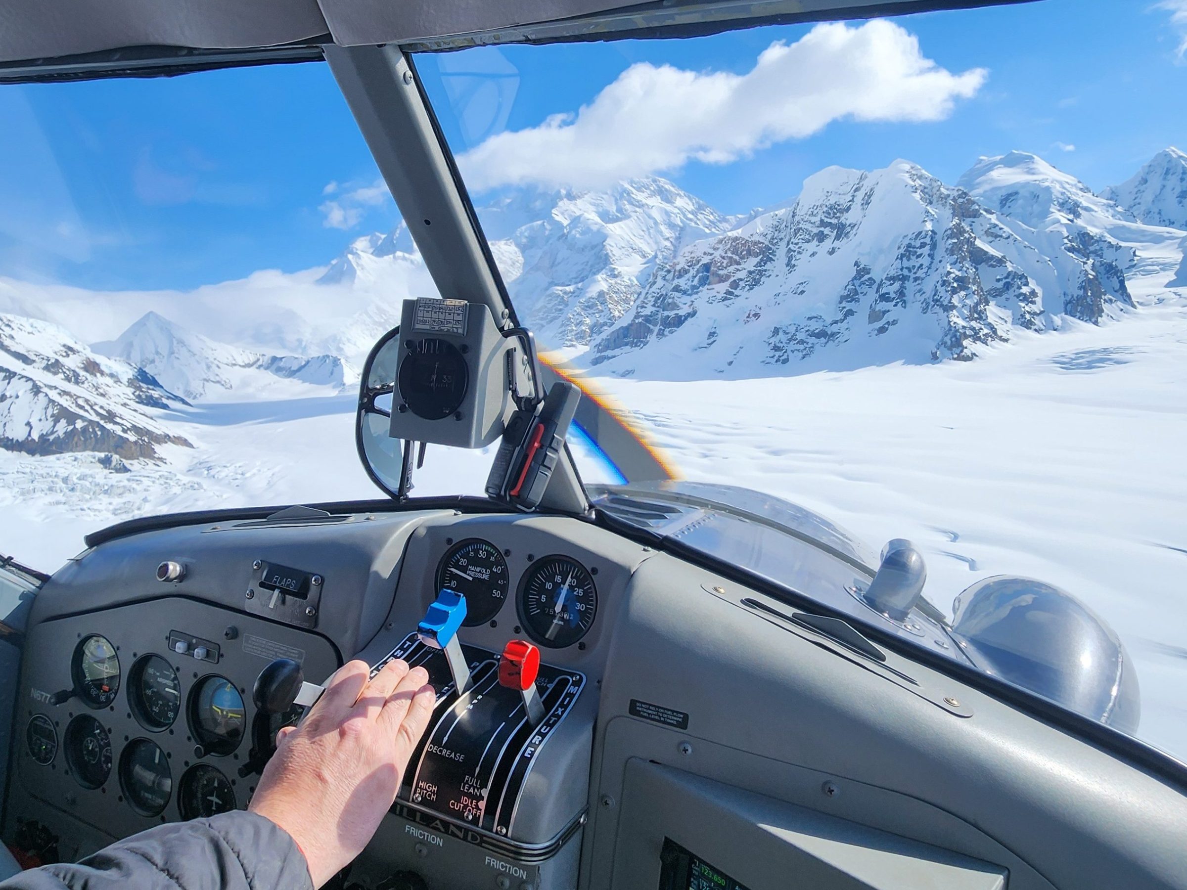 View from cockpit flying over snow-covered mountains under a clear blue sky.