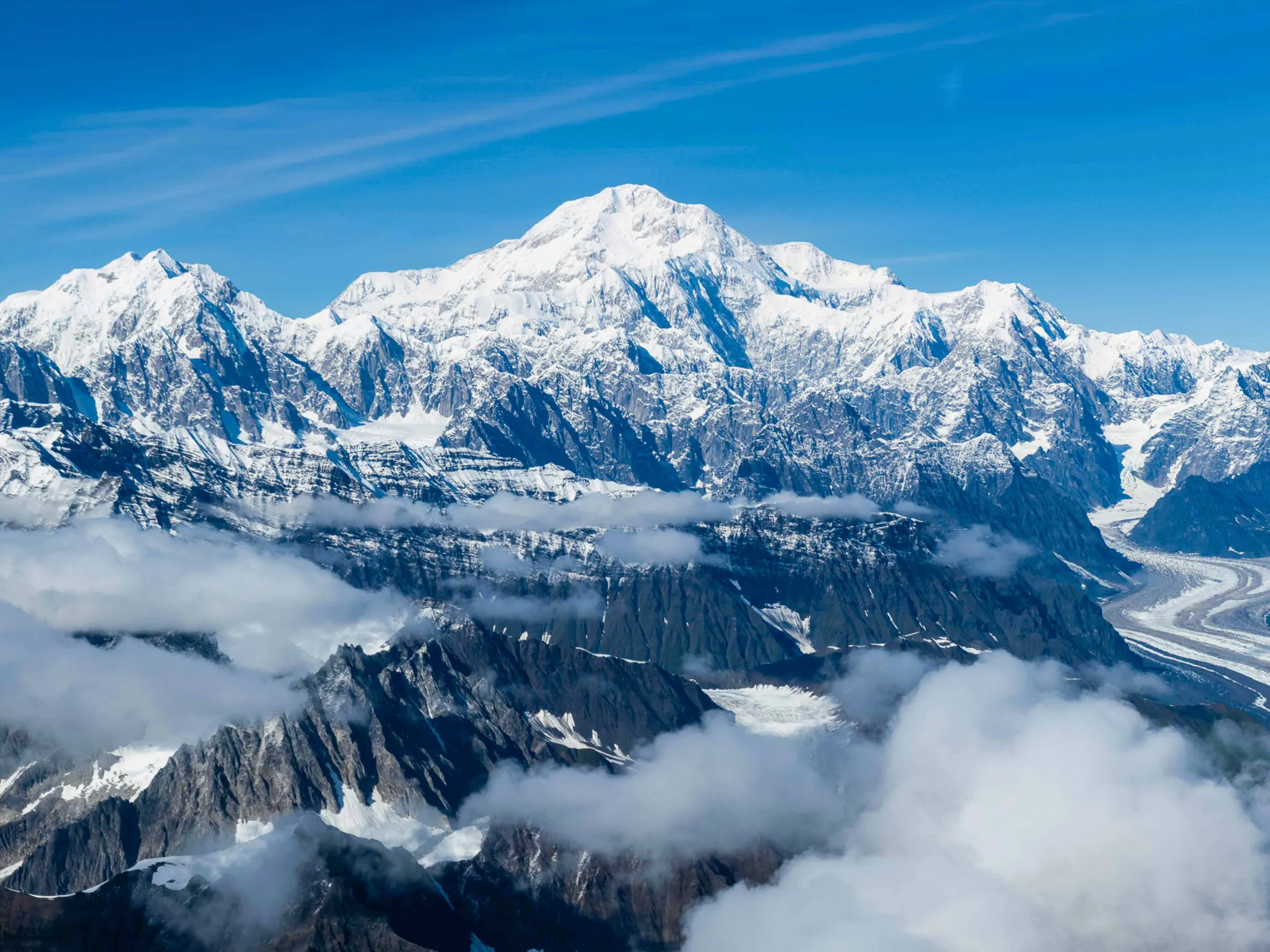 Snow-capped mountain range with clouds and a winding glacier under a clear blue sky.