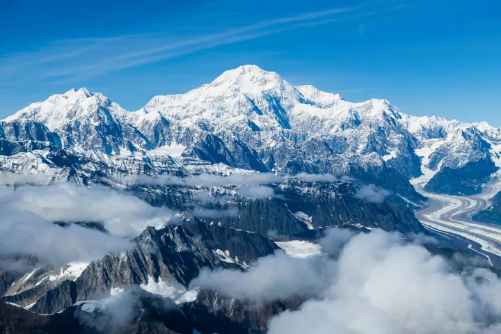 Snow-capped mountain range with clouds and a winding glacier under a clear blue sky.