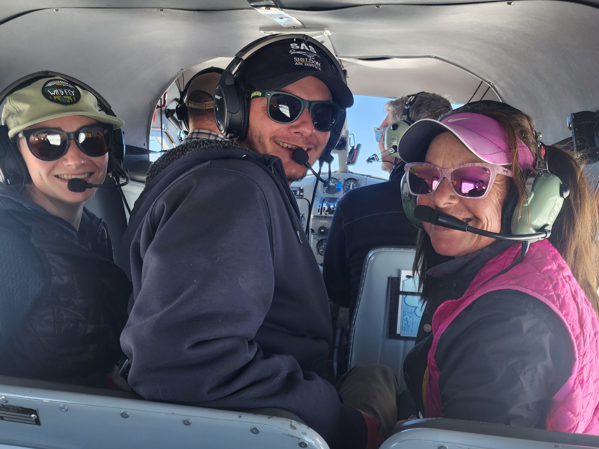 Three people with headsets in a small plane cabin with mountains visible outside.