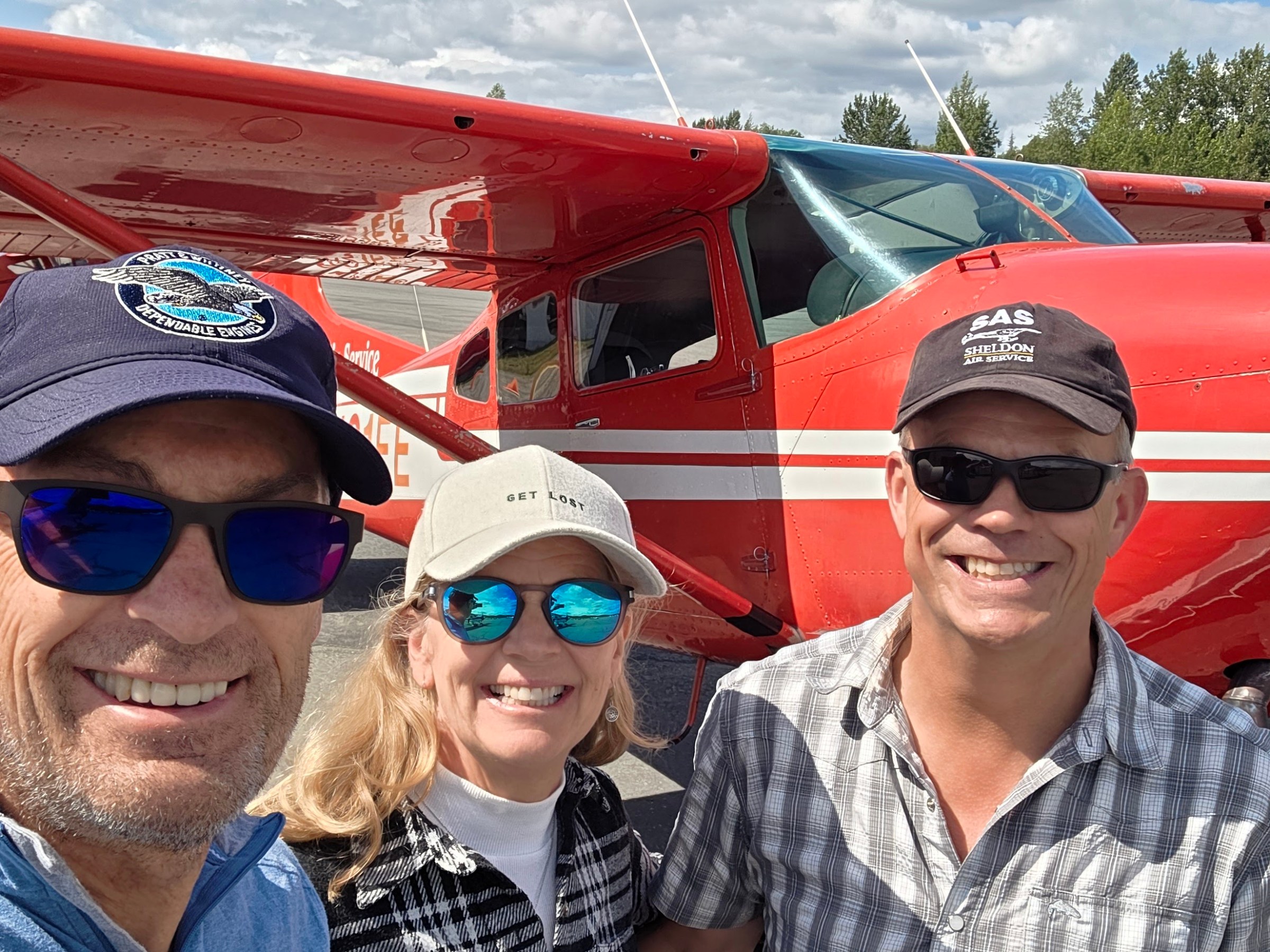Three people smiling in front of a red propeller airplane on a sunny day.