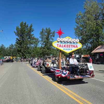 Parade with a float featuring 'Welcome to Talkeetna, Alaska' sign and musician playing keyboard, surrounded by spectators in Talkeetna Alaska