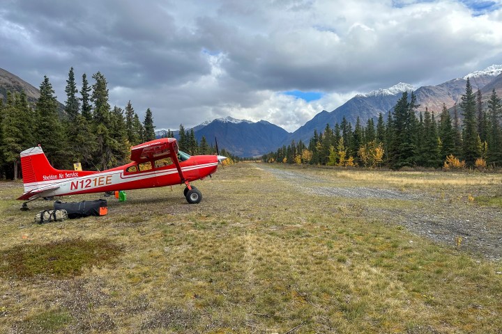 Red plane parked on grassy field near forest and mountains under cloudy sky.