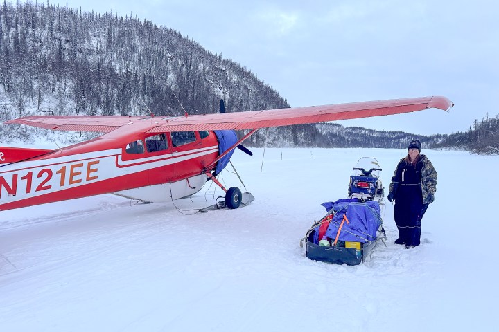 Red and white plane on snowy ground, person standing nearby with supplies in winter gear.