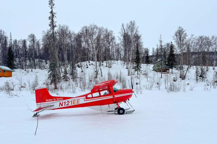 Red plane on snow with a background of leafless trees and wooden cabins.