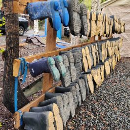 Rows of boots drying on a wooden rack outdoors near a tent and vehicle.