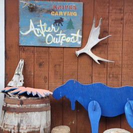 Rustic sign and wooden moose cutout on a wooden building with antler and barrel decor.