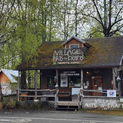 Rustic wooden craft shop surrounded by trees, with signs reading 'Village Arts and Crafts'.