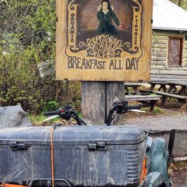 Wooden sign for Conscious Coffee above a parked ATV, with greenery and a log cabin in the background.