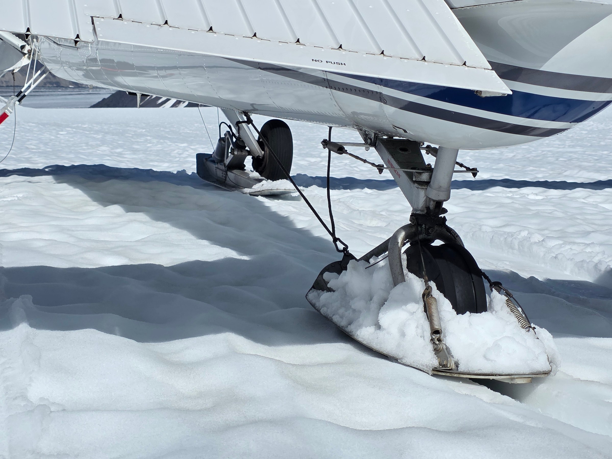 Close-up of airplane ski landing gear on snow-covered ground in bright sunlight.