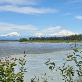 Snow-capped mountains and forest reflected in a calm river under a blue sky.