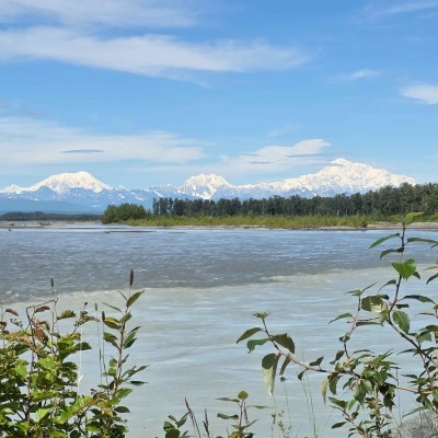Snow-capped mountains and forest reflected in a calm river under a blue sky.