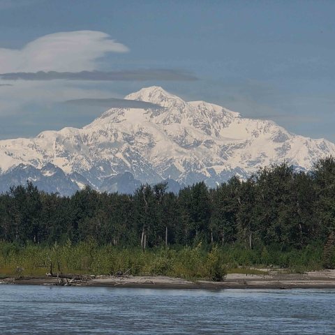 Snow-capped mountain with forest and river in foreground under a clear sky.