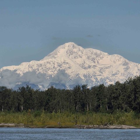 Snow-covered mountain with forest and river in the foreground under a clear sky.