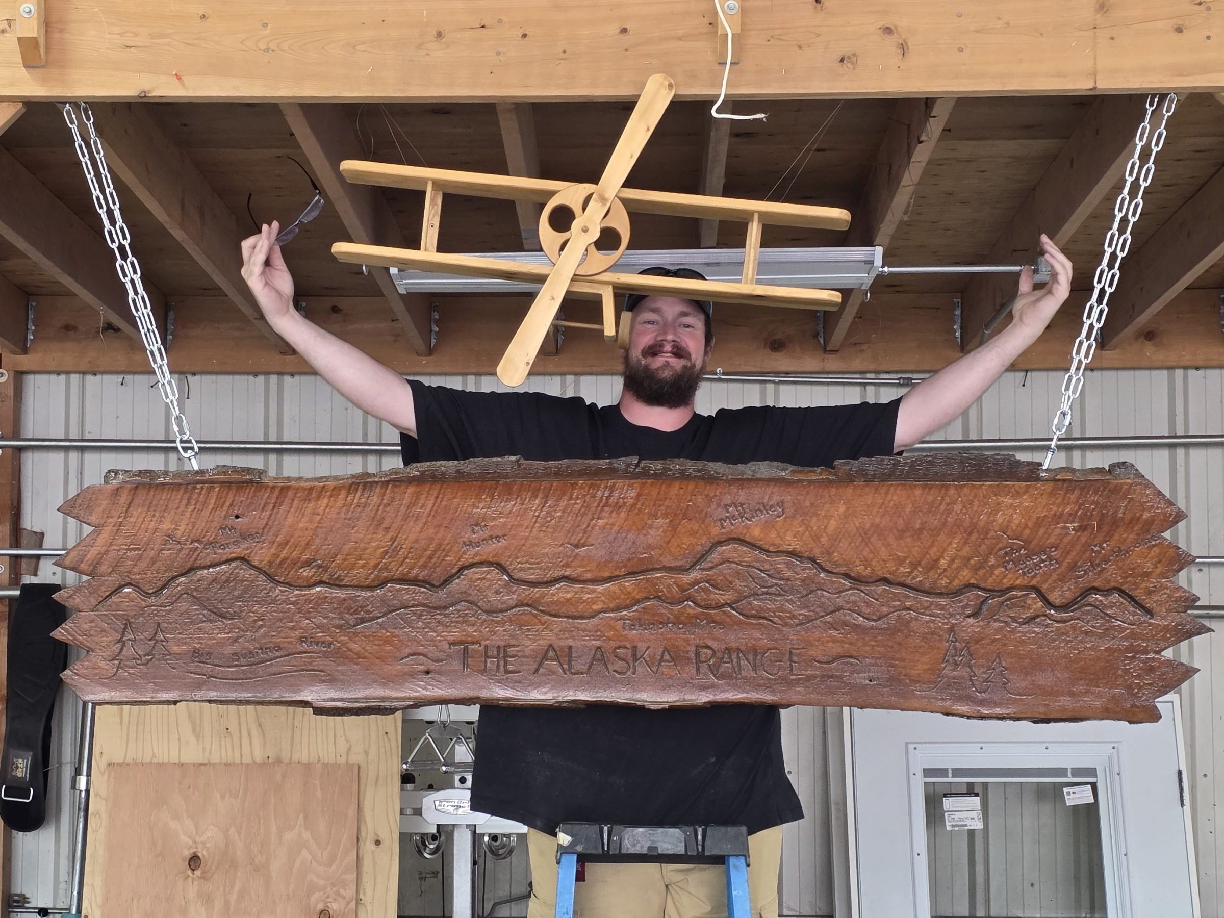Robert Stumpf ladder holding a wooden sign with 'The Alaska Range' at the Sheldon Hangar 2025