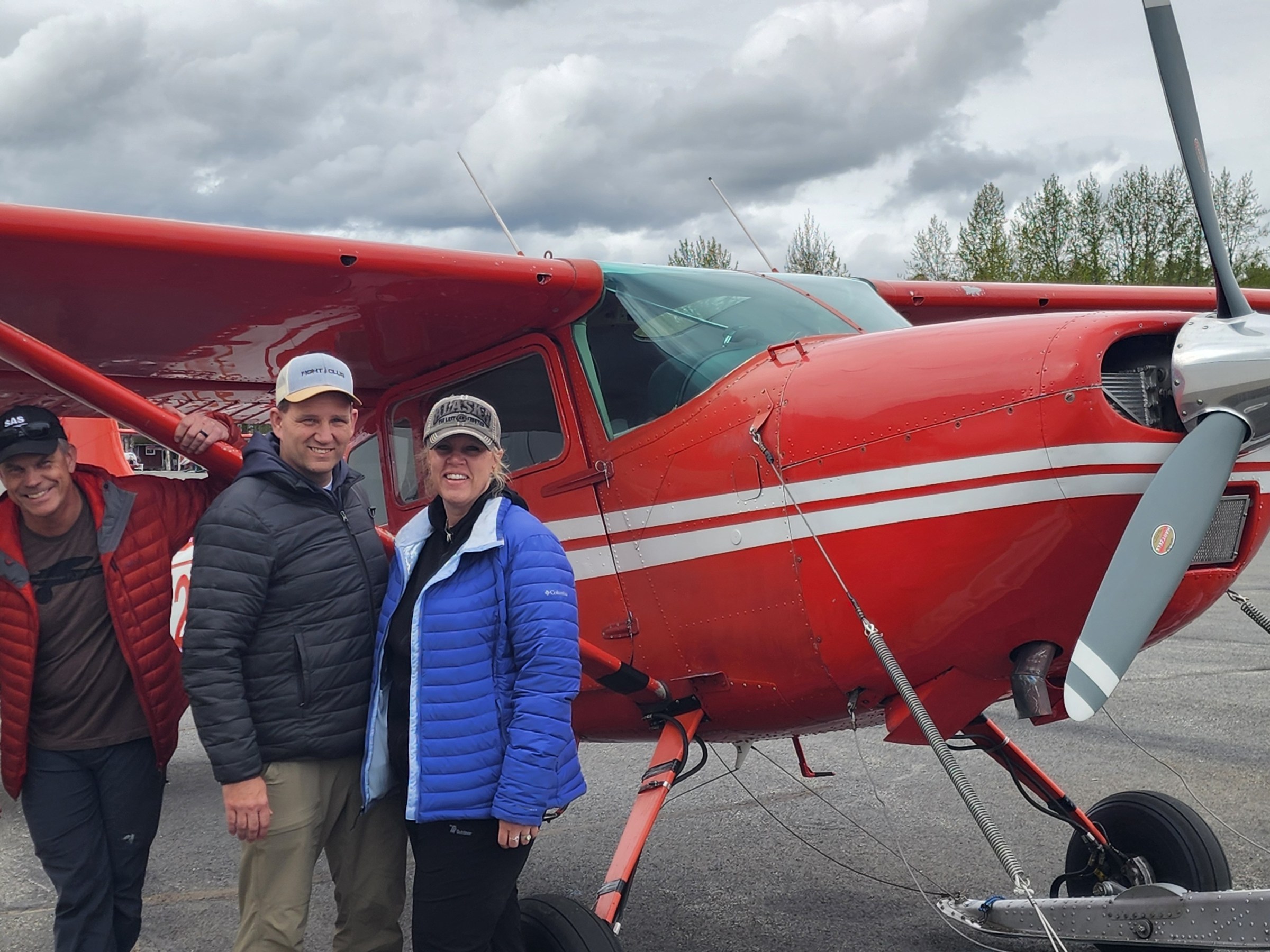 Three people in jackets standing by a small red airplane on a cloudy day.