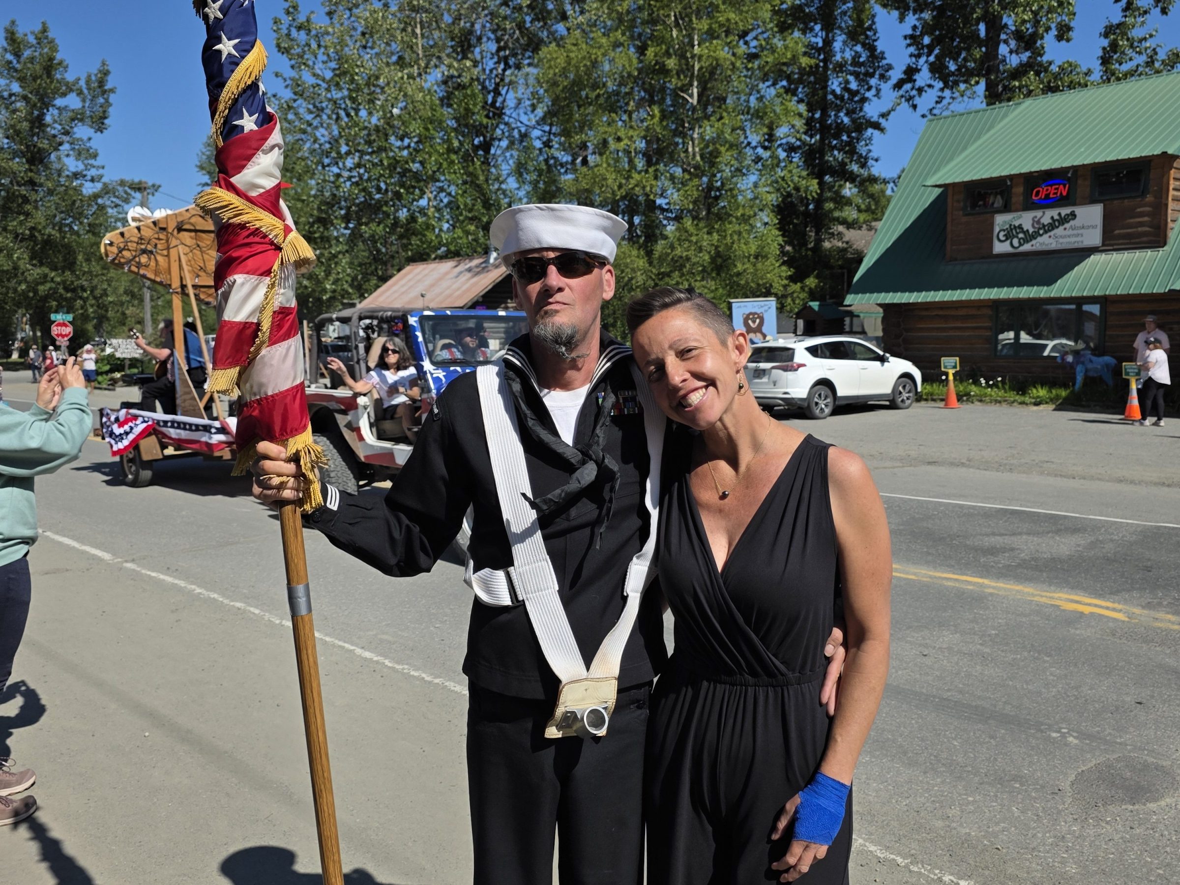 Two people pose next to each other, one holding a flag, outside with trees and a building in the background.