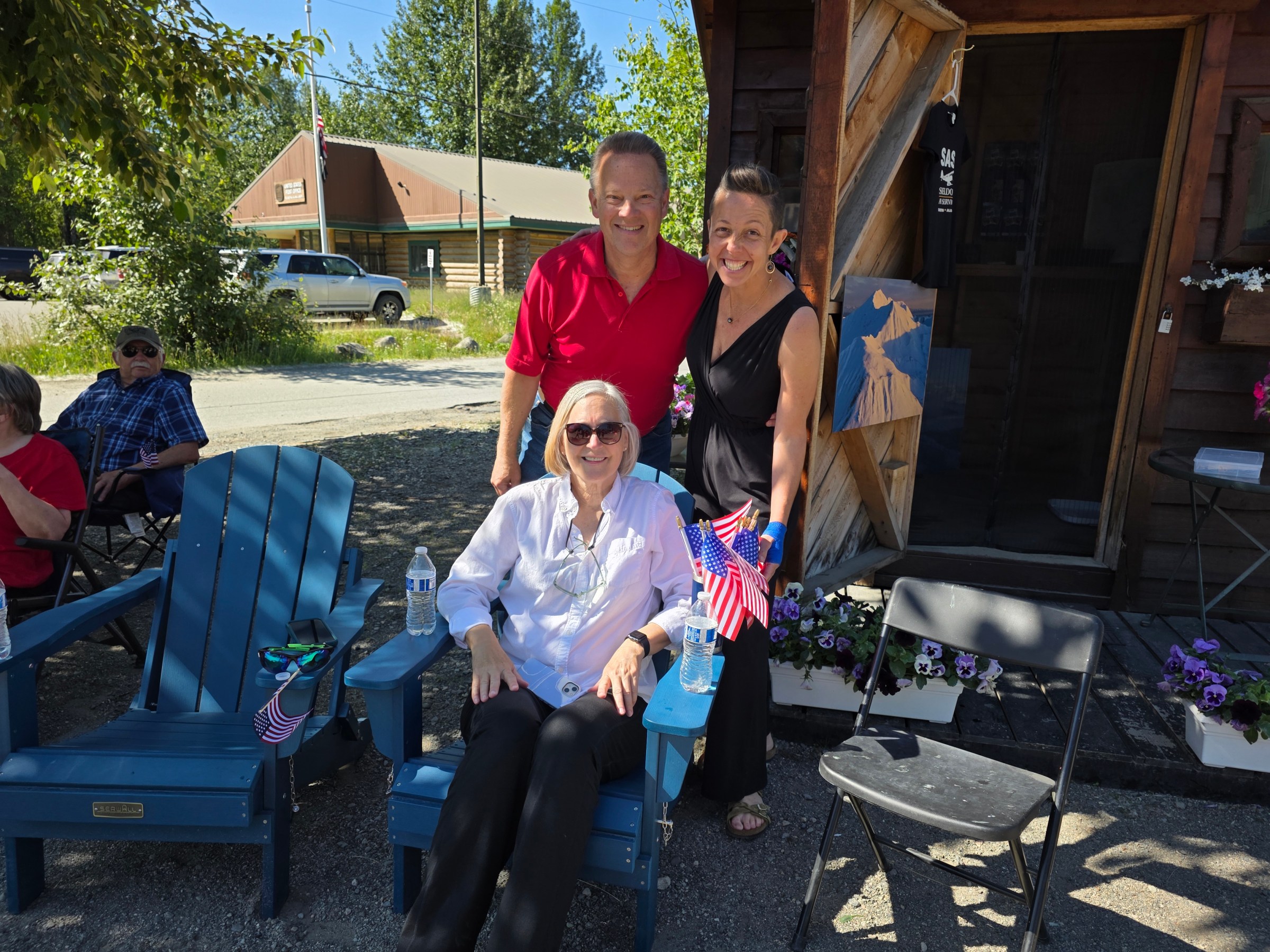 Three people pose outdoors, one seated holding small American flags, two standing behind, with houses and trees in background.