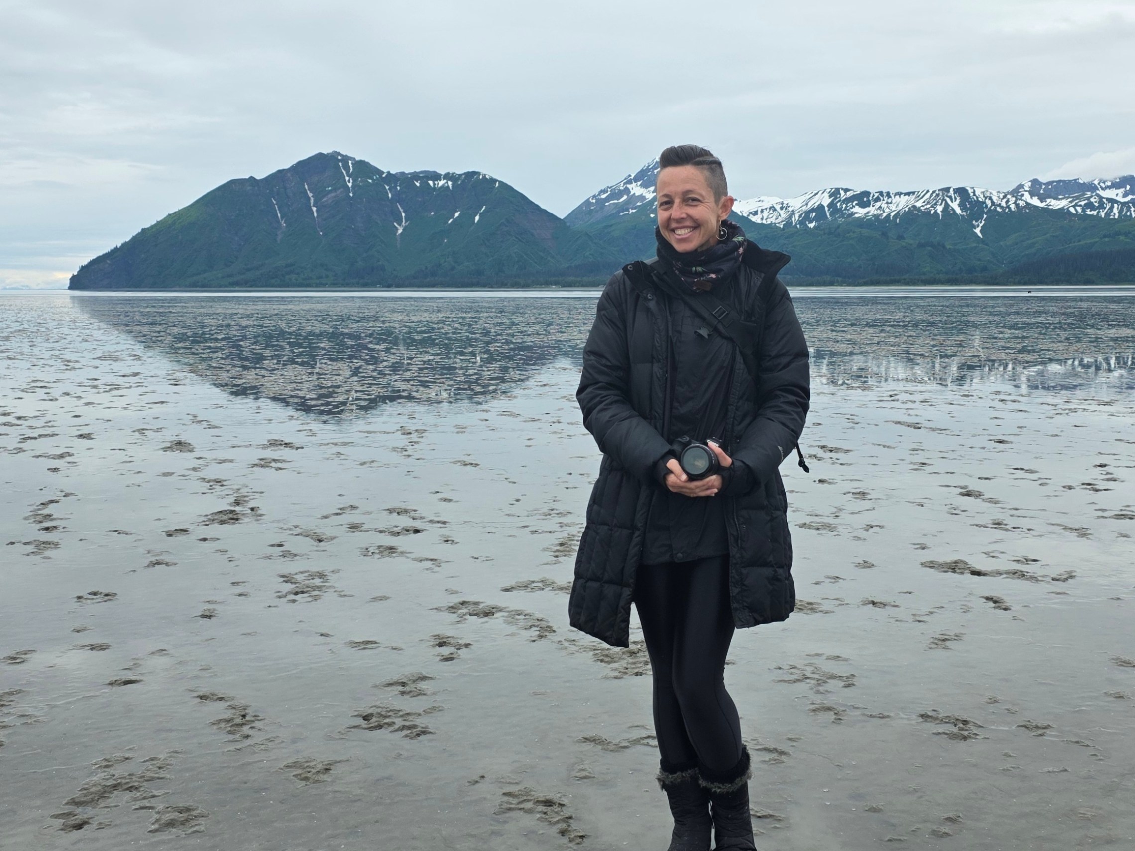 Person in black coat standing on wet sand with snowy mountains in the background.