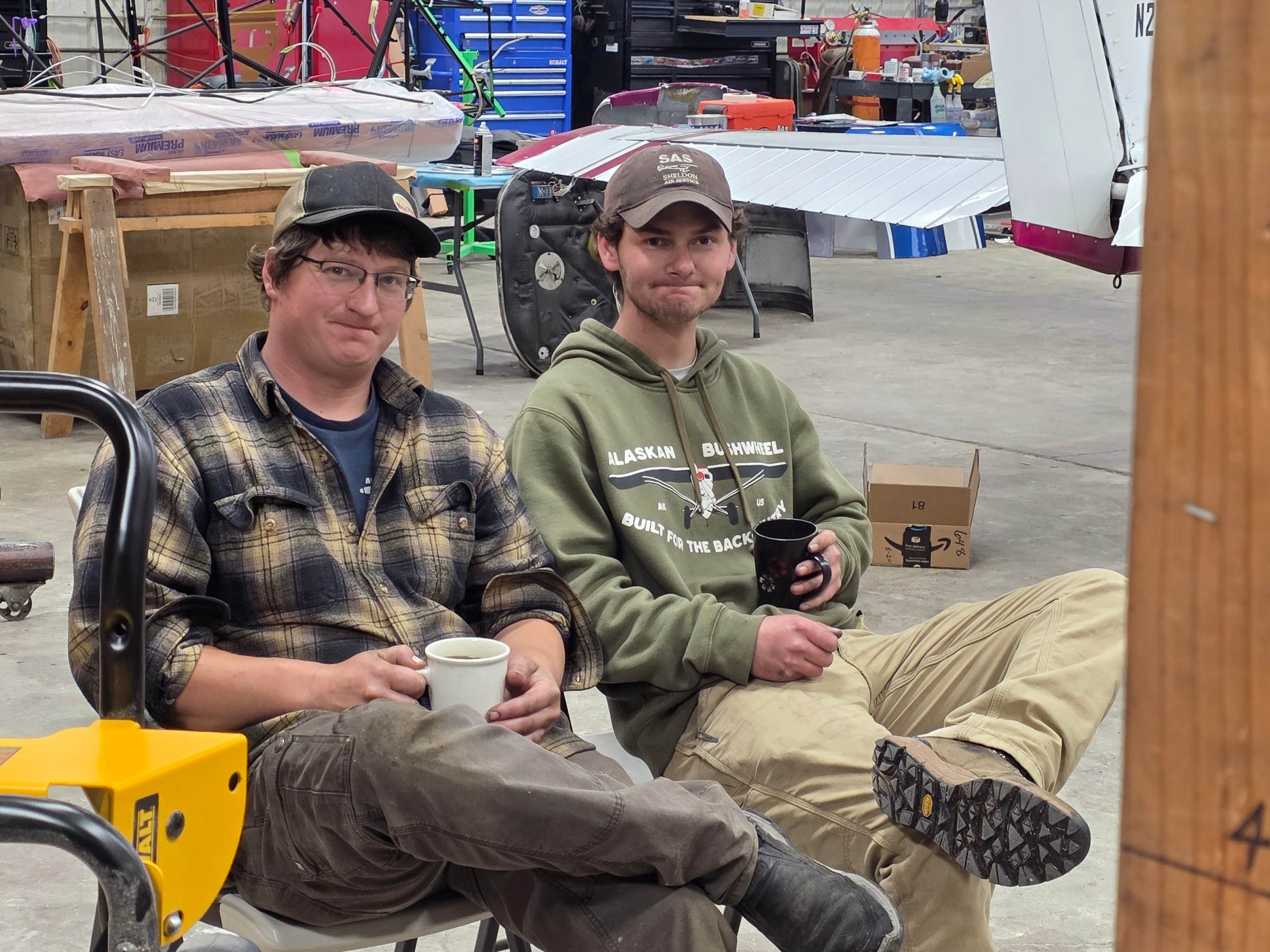 Two people sitting in a workshop, holding mugs, with airplane parts and tools in the background.