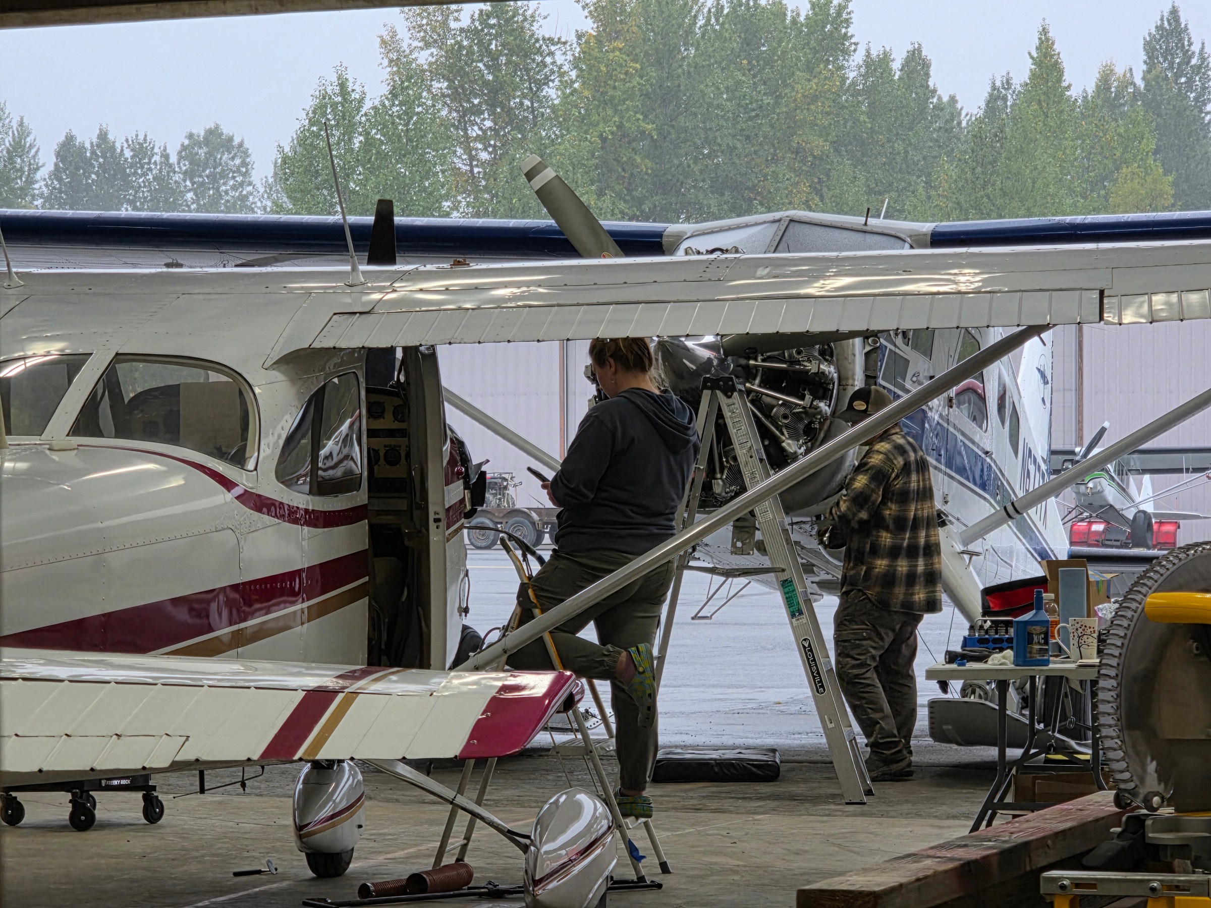 Two people working on small airplanes in a hangar with tools and ladders nearby.