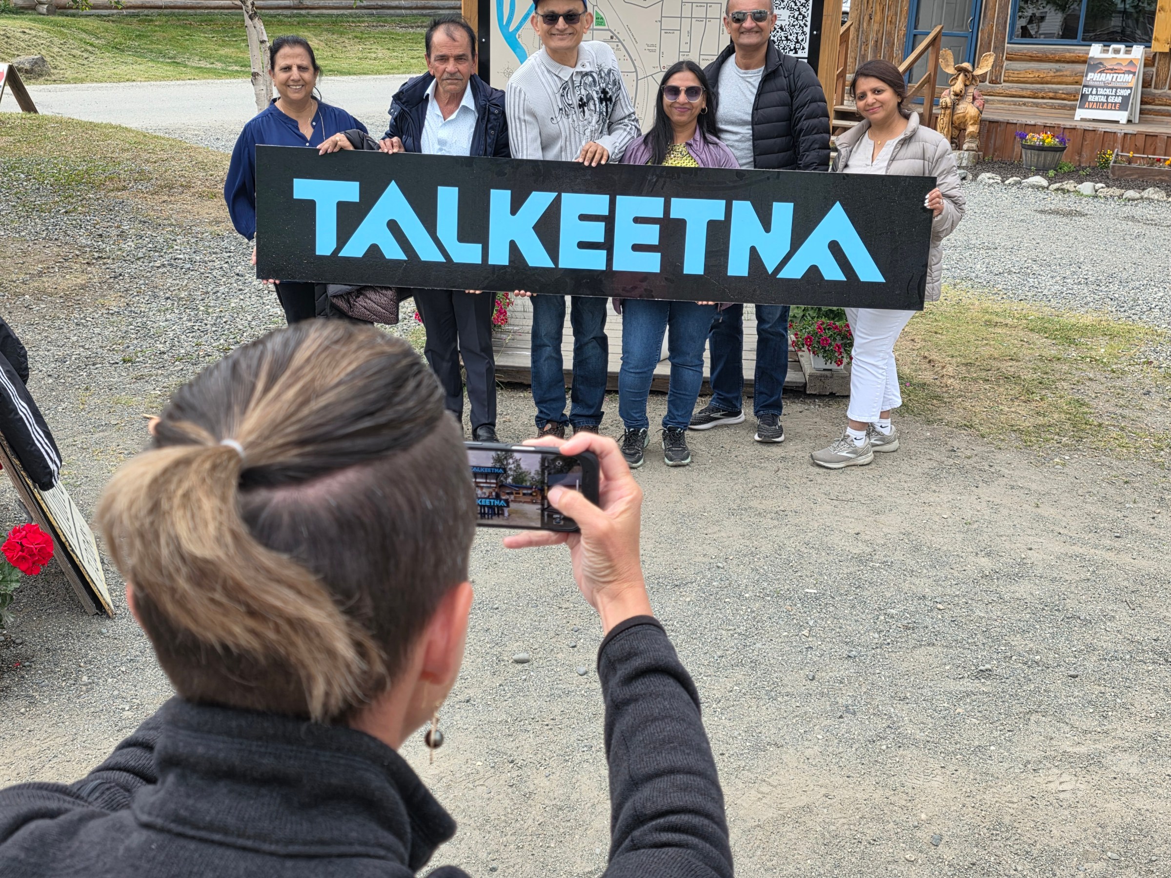 Person takes photo of six people holding 'TALKEETNA' sign outdoors.