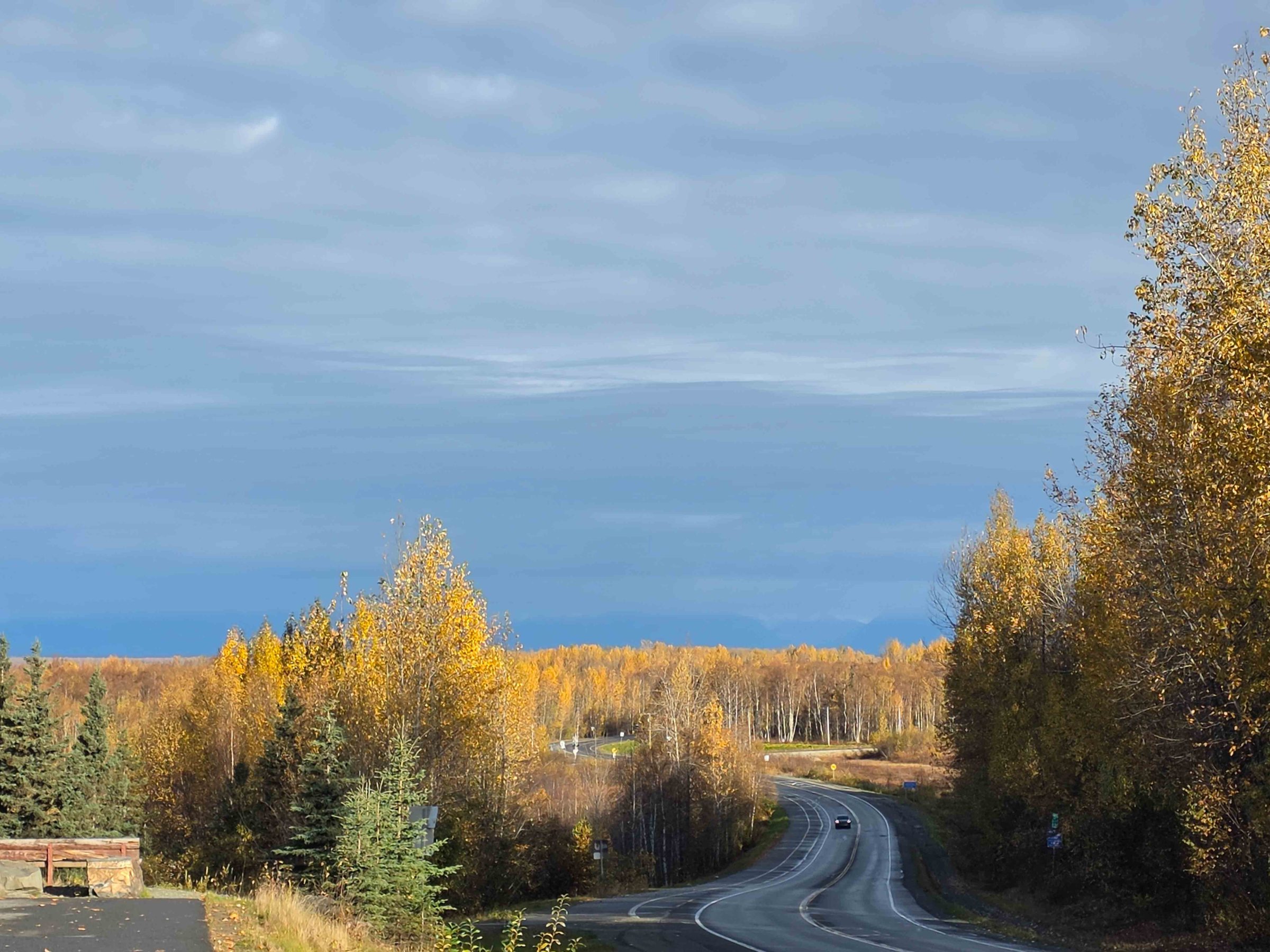 Curved road through autumn trees under cloudy sky, with two cars visible.