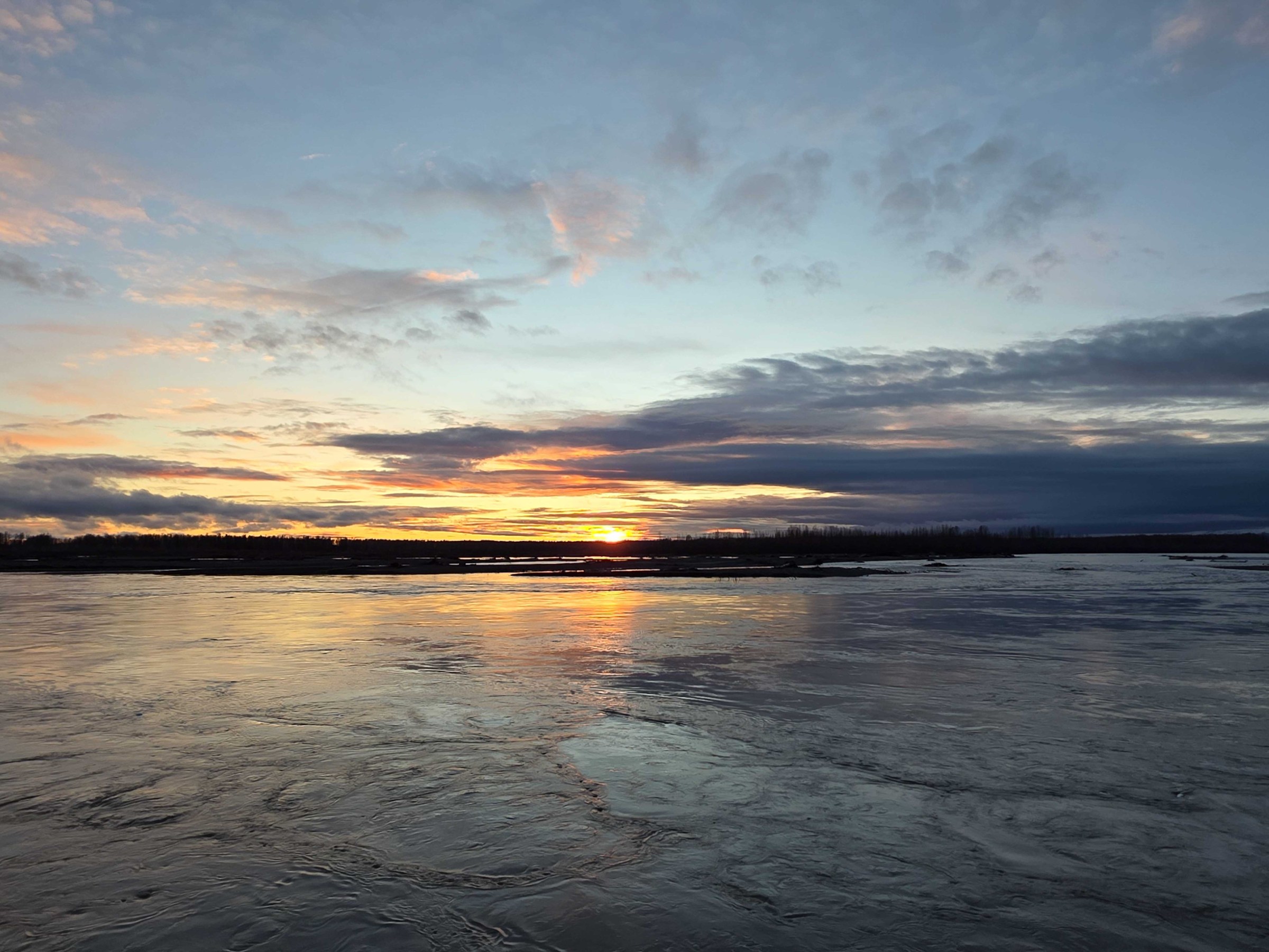 Sunset over a calm river with reflections and scattered clouds in the sky.