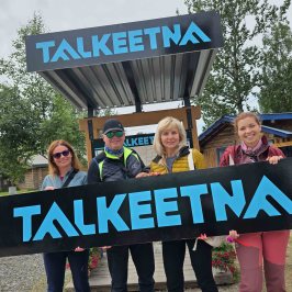 Four people hold a sign that says 'Talkeetna' with a building and trees in the background.