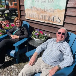 Two people sitting on blue chairs outside, smiling in sunny weather.