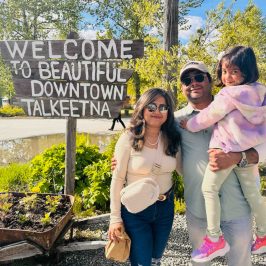 Family poses by 'Welcome to Downtown Talkeetna' sign with trees and sky in background.