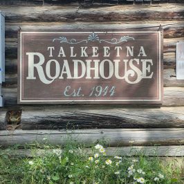 Wooden sign reading 'Talkeetna Roadhouse Est. 1944' with daisies below.