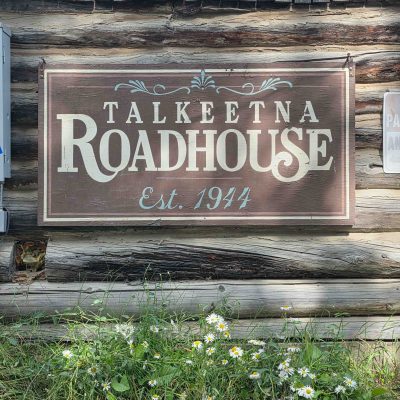 Wooden sign reading 'Talkeetna Roadhouse Est. 1944' with daisies below.