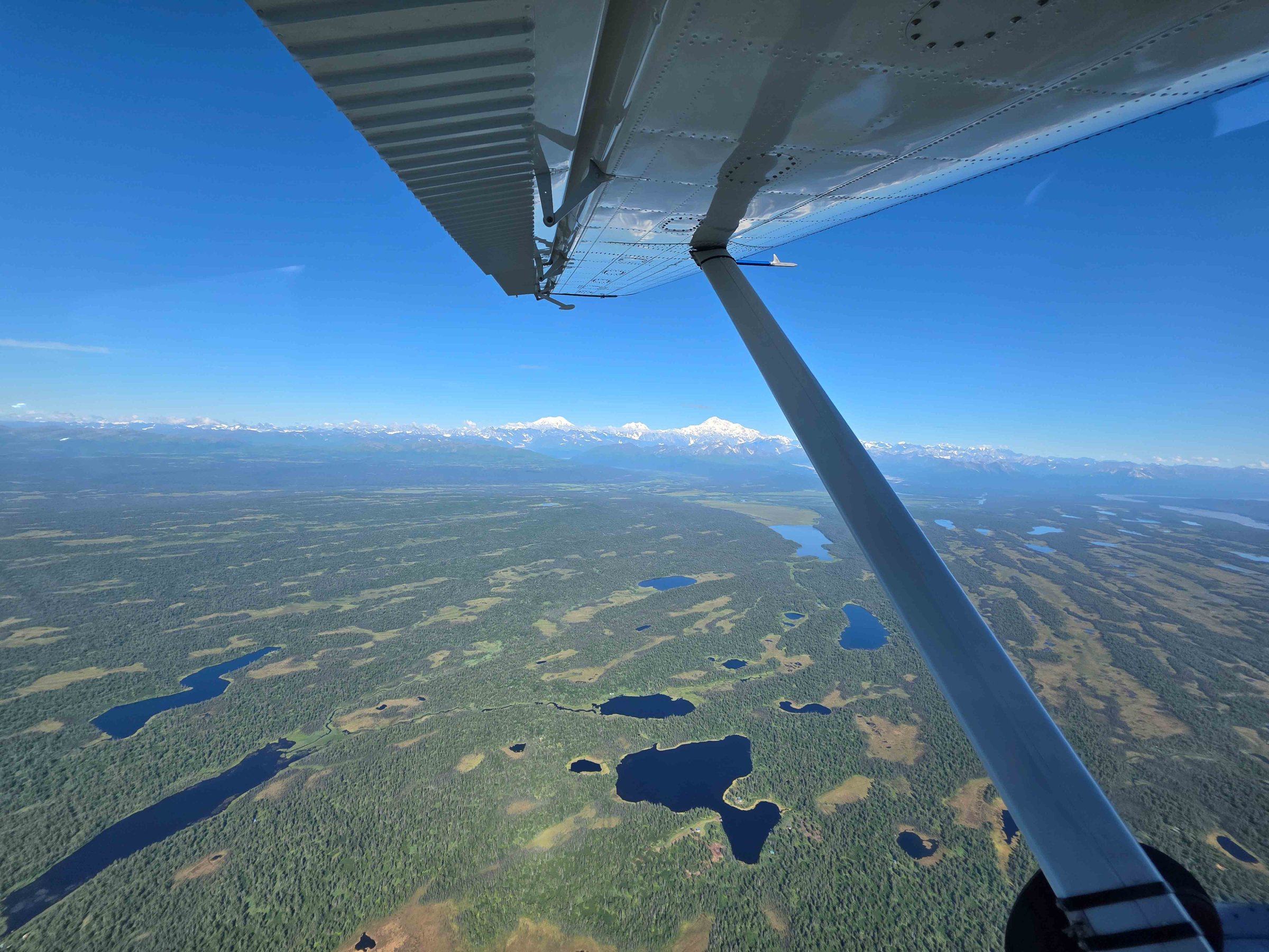 View from plane wing of vast landscape with lakes and mountains in the distance under clear blue sky.
