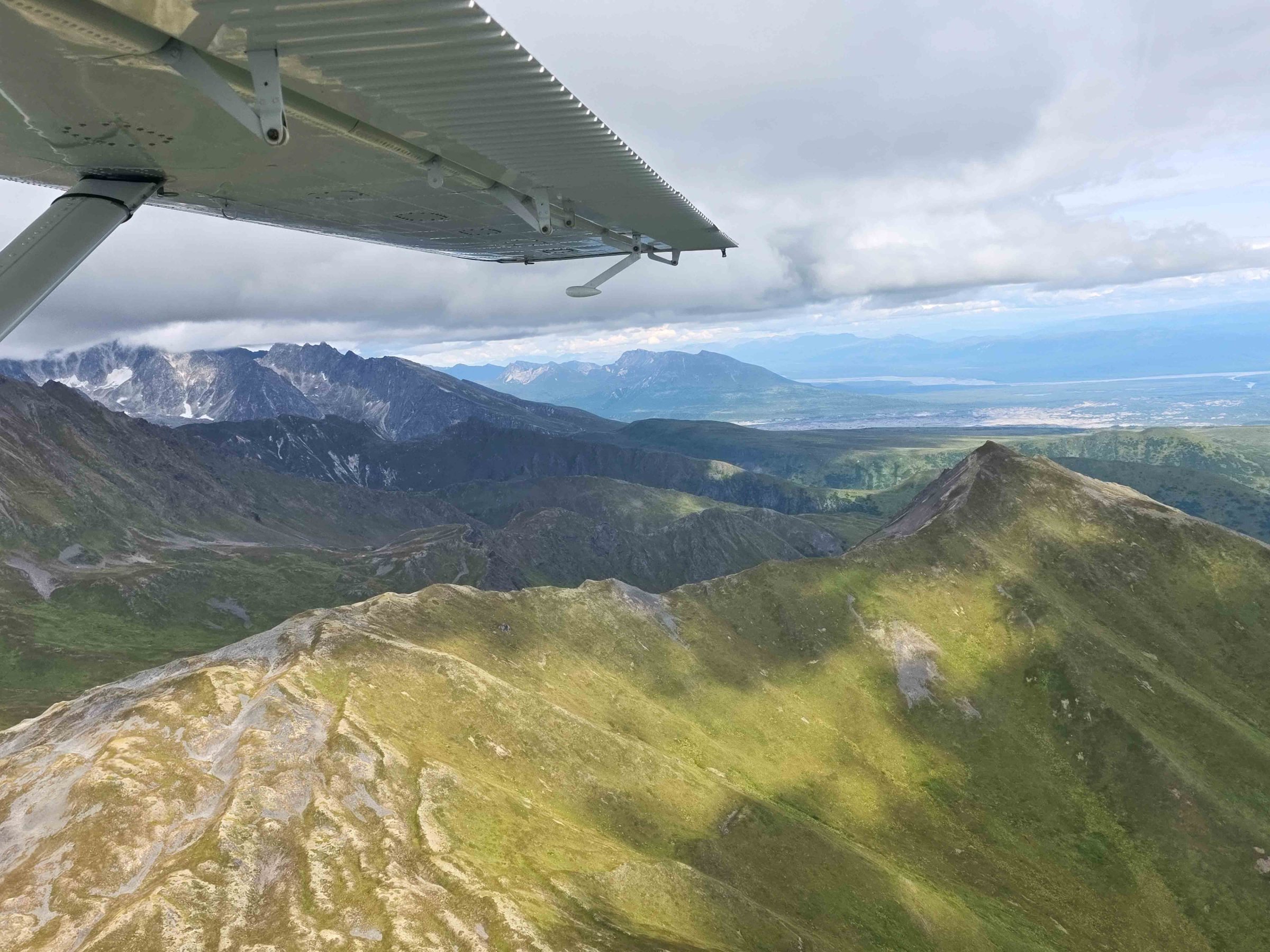 View from plane wing over rugged green mountains below cloudy sky.