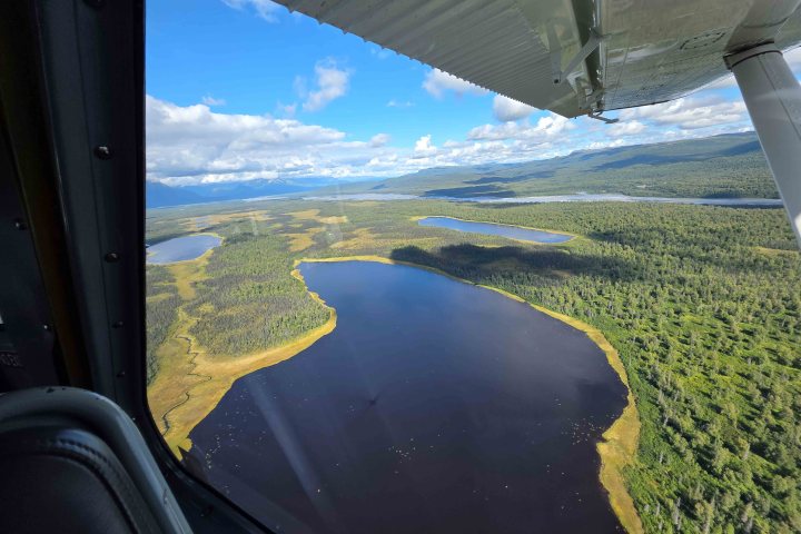 Aerial view from airplane window of a lake surrounded by forest and mountains under a cloudy blue sky.