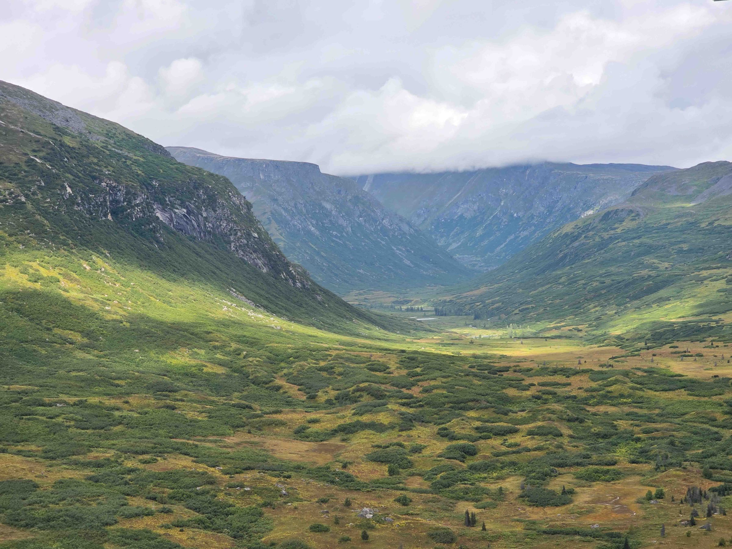 Cloudy mountain valley with green hills and a distant view of fog and peaks.