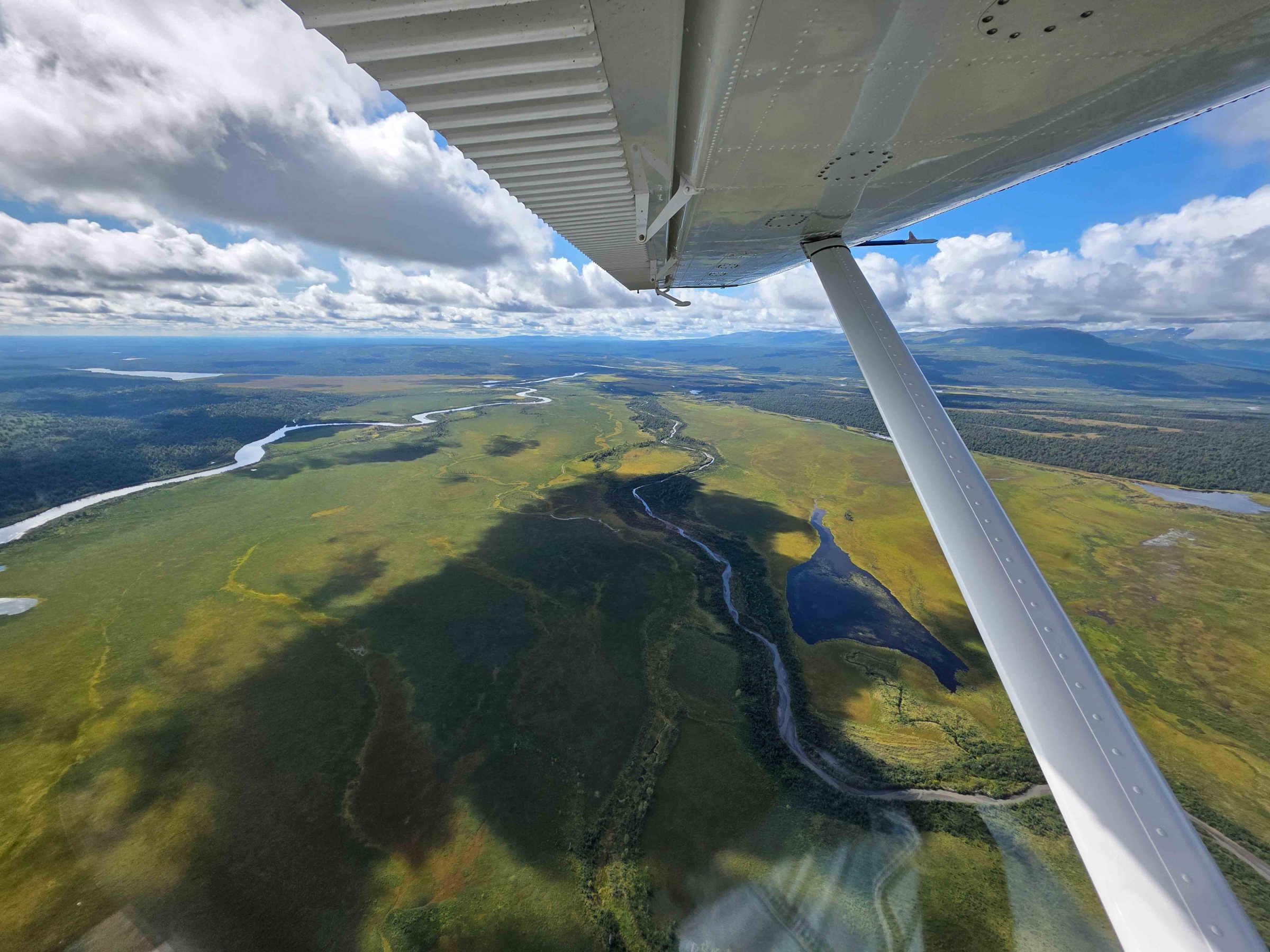 Airplane wing over vast green landscape with winding rivers and cloudy sky.
