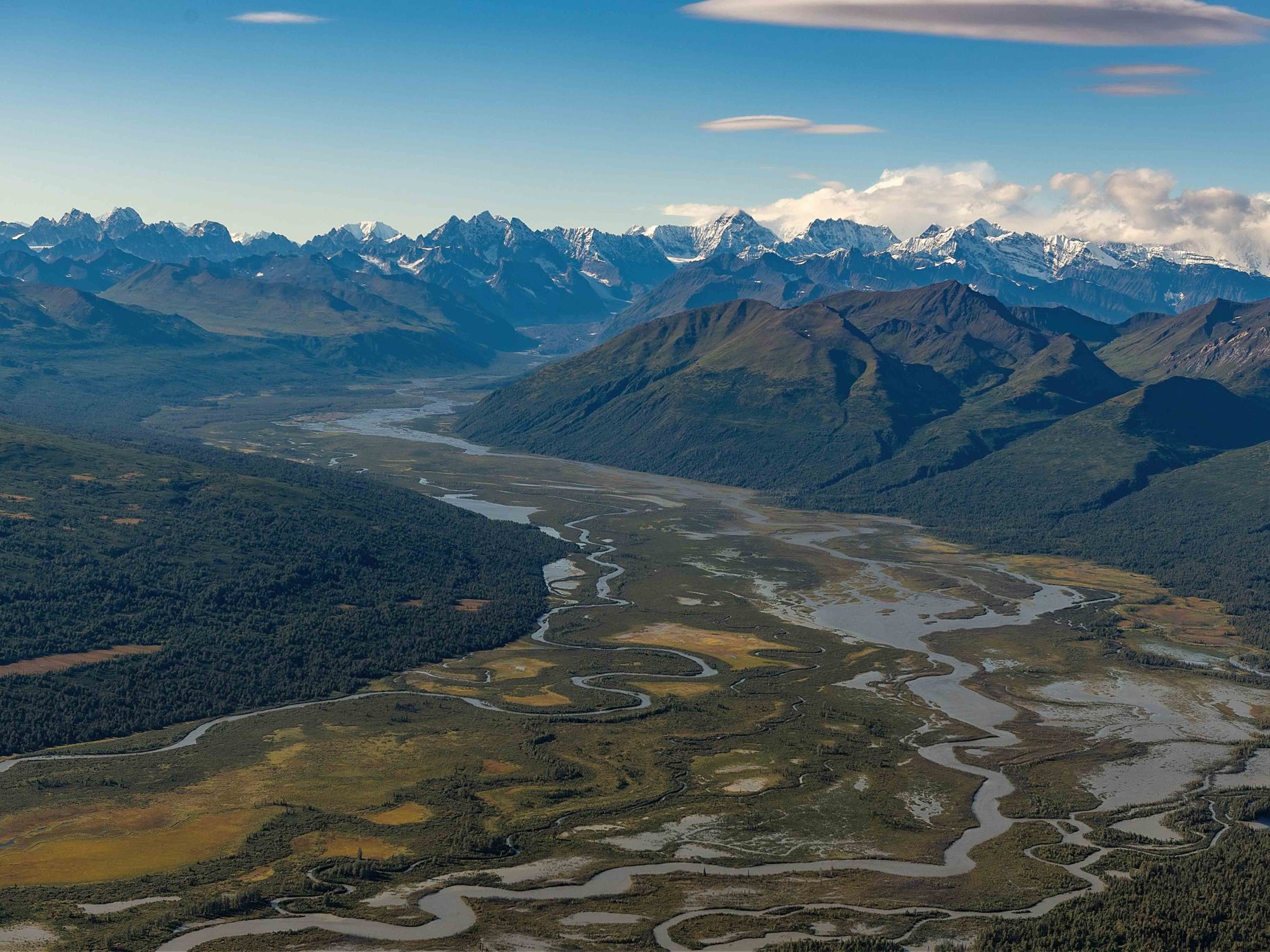Aerial view of winding river valley with snow-capped mountains in the background.