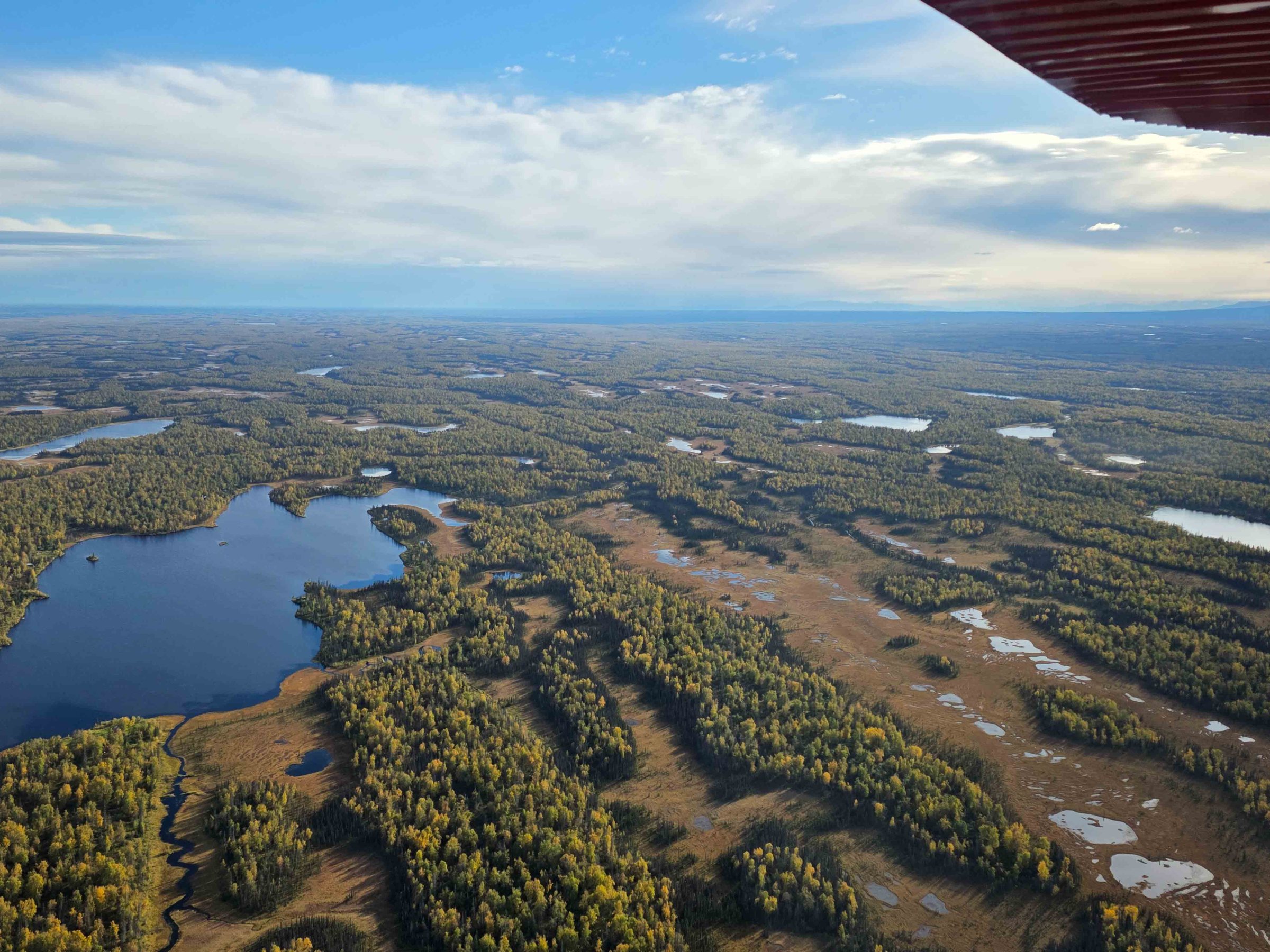Aerial view of a vast landscape with lakes, forests, and distant mountains under a partly cloudy sky.