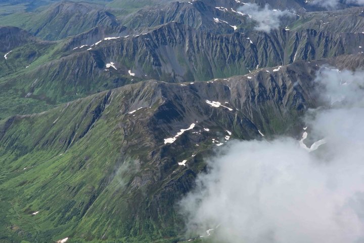 Aerial view of green mountains with patches of snow and clouds.
