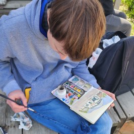 Miles painting outdoors on a sketchbook, nearby another holding Aurora the mayor