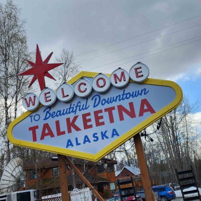 Vegas Style Welcome sign for Talkeetna, Alaska, with trees and cloudy sky background.