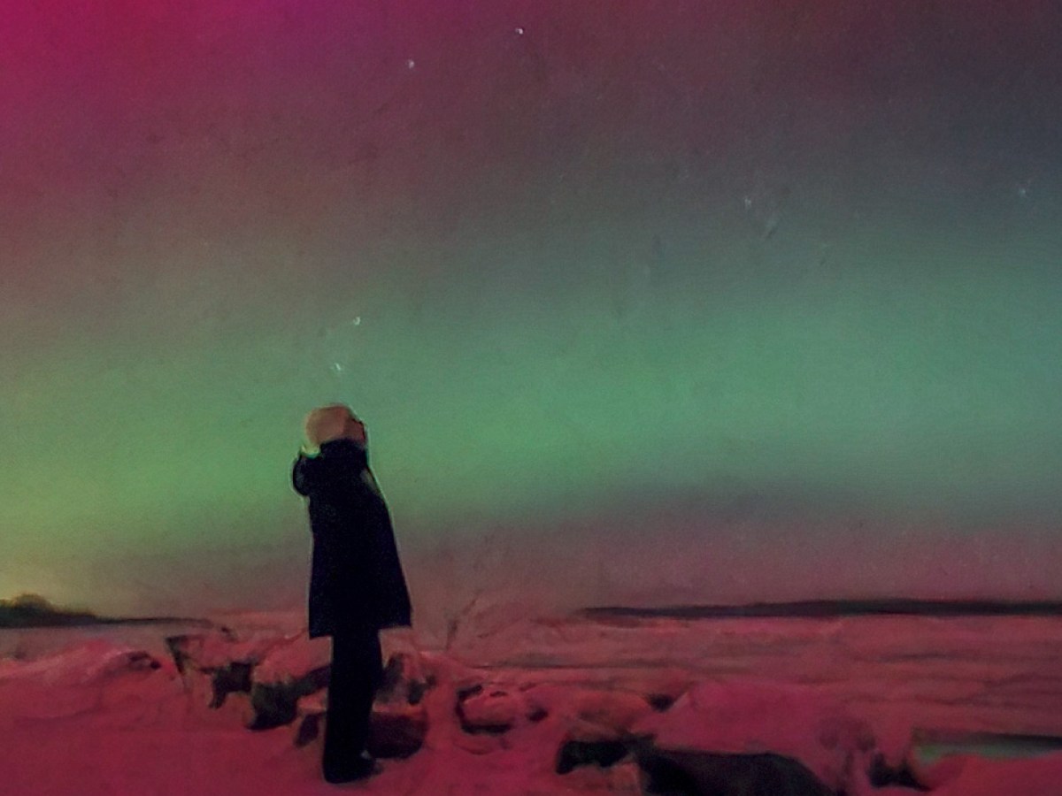 Person standing on snowy ground under a vibrant aurora with red and green lights in the night sky by the Susitna River in Alaska