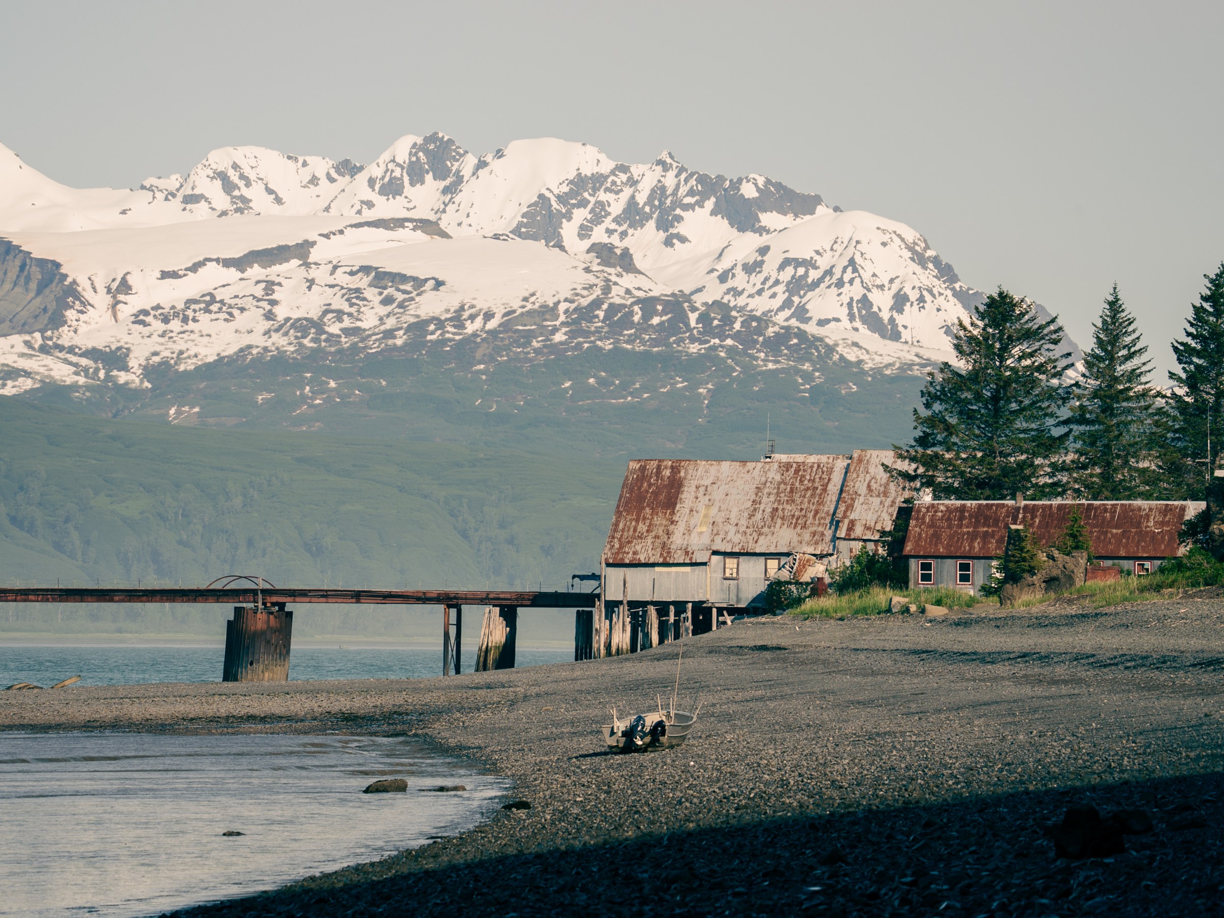 Rustic buildings by a beach with a pier, snow-capped mountains in the background.