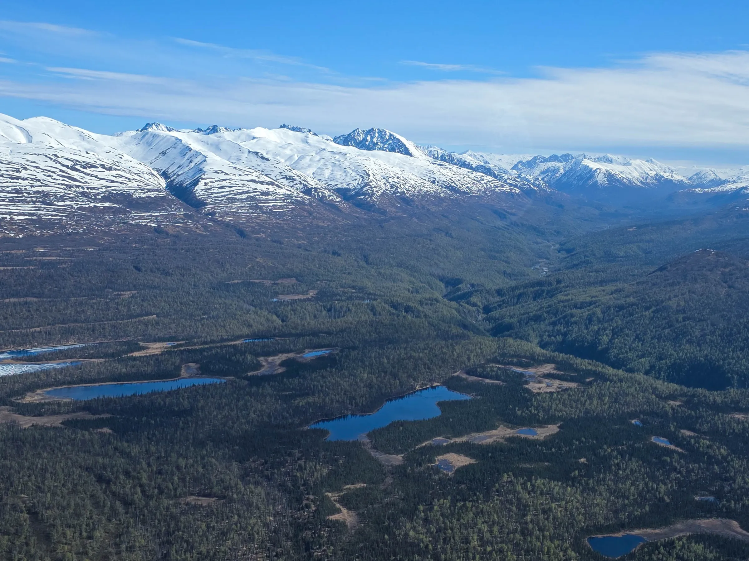 Aerial view of snow-capped mountains and forests with small lakes under a blue sky.