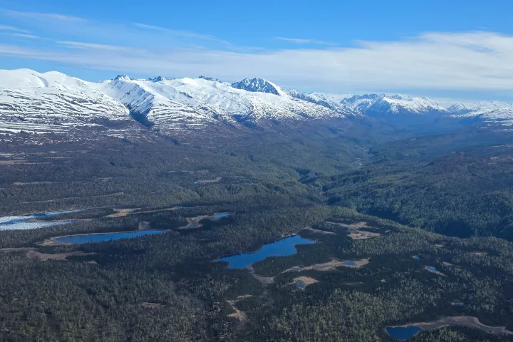 Aerial view of snow-capped mountains and forests with small lakes under a blue sky.